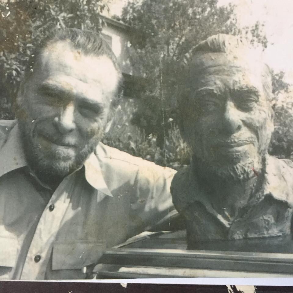 Old black and white photo of Charles Bukowski smiling next to a sculpted bust of his own head
