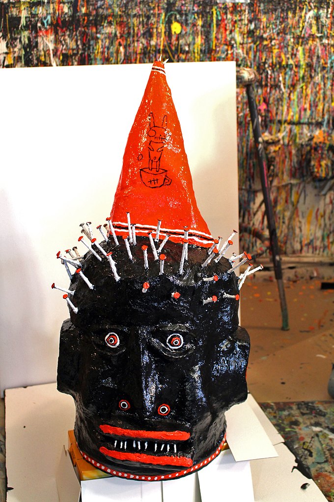 A black painted head made from a tin, with red eyes and lips, white teeth, a ring of nails and a tall orange dunce cap, in front of a paint-splattered studio wall